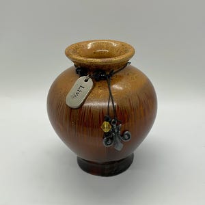 May include: A round ceramic vase with a glossy brown and tan drip glaze finish. A black cord is tied around the neck, featuring a silver-toned metal tag engraved with the word "Live," a small yellow crystal bead, and a silver-toned metal fleur-de-lis charm.