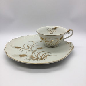 May include: A white porcelain teacup and saucer set with a gold wheat design. The cup has a handle and the saucer has a scalloped edge.