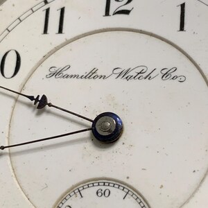 Op de afbeelding: Close-up van een vintage horlogekast. De witte wijzerplaat heeft zwarte cijfers, een kleinere secondenwijzerplaat en de inscriptie "Hamilton Watch Co.". De wijzers zijn zwart en de minutenmarkeringen rood.
