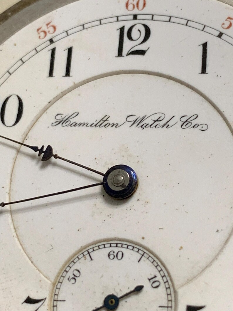 Op de afbeelding: Close-up van een vintage horlogekast. De witte wijzerplaat heeft zwarte cijfers, een kleinere secondenwijzerplaat en de inscriptie "Hamilton Watch Co.". De wijzers zijn zwart en de minutenmarkeringen rood.