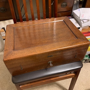 May include: A wooden box with a keyhole and a drawer. The box is sitting on a wooden chair with a black leather seat.
