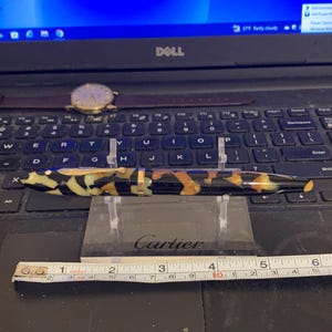 May include: A black and gold Cartier pen with a tortoiseshell-like pattern. The pen is resting on a clear acrylic stand. A ruler is in the foreground.