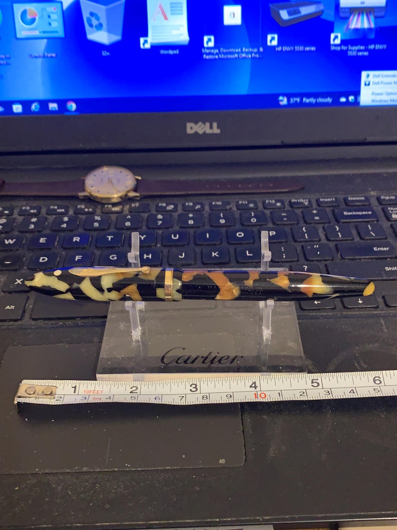 May include: A black and gold Cartier pen with a tortoiseshell-like pattern. The pen is resting on a clear acrylic stand. A ruler is in the foreground.
