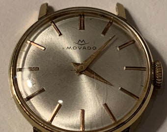 May include: A gold-toned wristwatch with a white face and black hands. The watch face has a black logo that says "MOVADO" with a crown above the word. The watch has a gold-toned band.