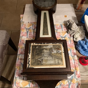 May include: A vintage wooden clock with a Roman numeral face and a picture of a bridge and a river in a frame below the clock face.