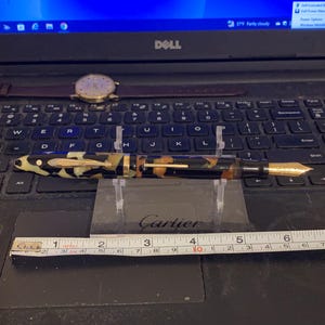 May include: A black and gold fountain pen with a tortoiseshell pattern on the barrel. The pen is resting on a ruler that reads "Cartier" and is marked in inches.