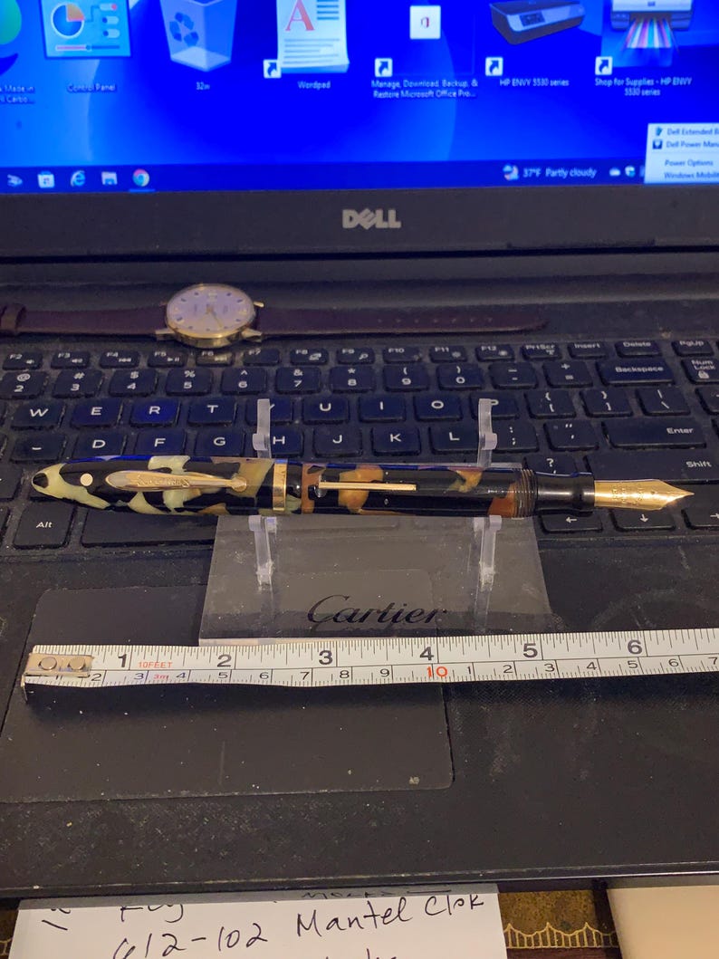 May include: A black and gold fountain pen with a tortoiseshell pattern on the barrel. The pen is resting on a ruler that reads "Cartier" and is marked in inches.