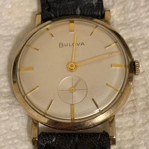 May include: A gold-toned Bulova wristwatch with a cream-colored face and gold hands and markers. The watch has a small seconds dial and a black leather band. The watch face displays the brand name "Bulova".