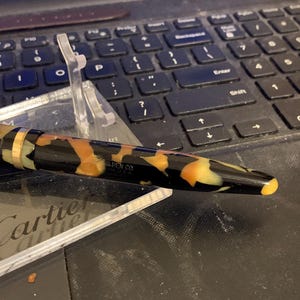 May include: A black and brown patterned fountain pen with gold accents. The pen is on a clear plastic stand and has the text "PEN CO. U.S.A." on the side.