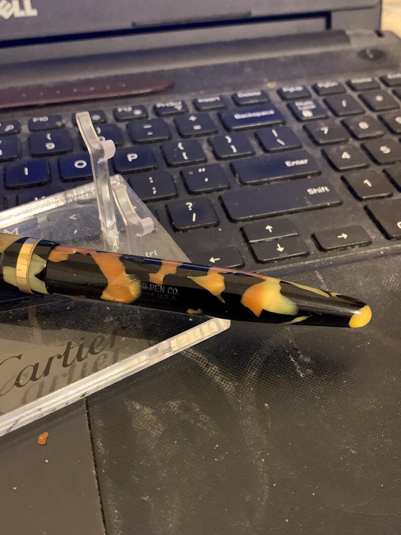 May include: A black and brown patterned fountain pen with gold accents. The pen is on a clear plastic stand and has the text "PEN CO. U.S.A." on the side.
