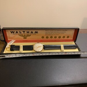 Op de afbeelding: Een gouden Waltham-horloge met een witte wijzerplaat en een zwart leren bandje in een zwarte doos met een gouden adelaarlogo en de tekst "Waltham Timing America for Over 100 Years".