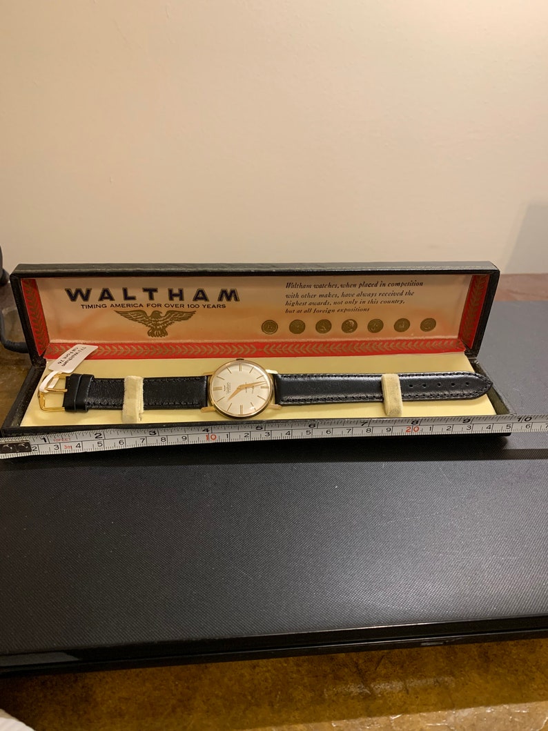 Op de afbeelding: Een gouden Waltham-horloge met een witte wijzerplaat en een zwart leren bandje in een zwarte doos met een gouden adelaarlogo en de tekst "Waltham Timing America for Over 100 Years".