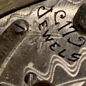 Op de afbeelding: Close-up van een vintage horlogekast met de inscriptie "17 JEWELS" in zwarte letters. De horlogekast heeft een textuur, patroonontwerp met een klein stervormig detail. Het kleurenschema is zilver en grijs.