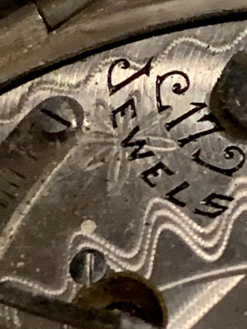Op de afbeelding: Close-up van een vintage horlogekast met de inscriptie "17 JEWELS" in zwarte letters. De horlogekast heeft een textuur, patroonontwerp met een klein stervormig detail. Het kleurenschema is zilver en grijs.