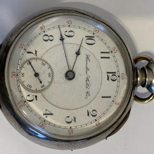 Op de afbeelding: Een zilveren Hamilton zakhorloge met een witte wijzerplaat, zwarte cijfers en een kleinere subwijzerplaat. Het horloge heeft een goudkleurige kroon en een secondewijzer. De woorden "Hamilton Watch Co." staan op de wijzerplaat.