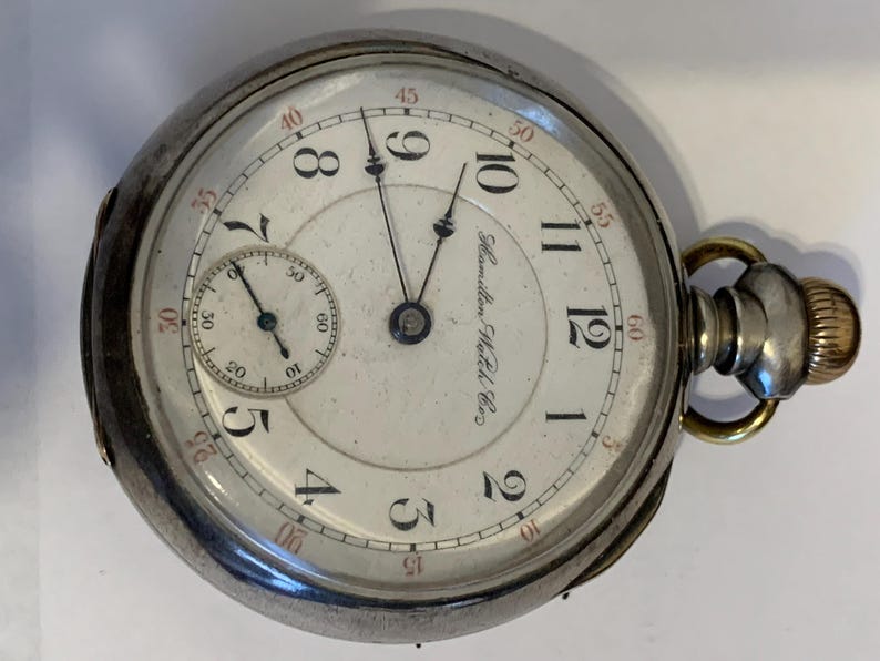 Op de afbeelding: Een zilveren Hamilton zakhorloge met een witte wijzerplaat, zwarte cijfers en een kleinere subwijzerplaat. Het horloge heeft een goudkleurige kroon en een secondewijzer. De woorden "Hamilton Watch Co." staan op de wijzerplaat.