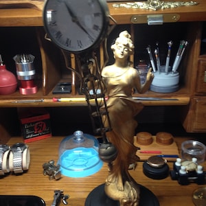 May include: A gold-toned clock with a Roman numeral face and a gold-toned figure of a woman standing on a black base. The clock is hanging from a chain and has a pendulum.