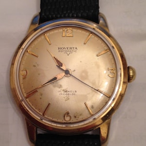 Puede incluir: Un reloj de pulsera dorado con una correa de cuero negro. La esfera del reloj es blanca con marcas negras y el texto "Hoverta Rotomatic 17 Jewels Incabloc".