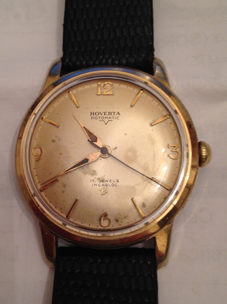 Puede incluir: Un reloj de pulsera dorado con una correa de cuero negro. La esfera del reloj es blanca con marcas negras y el texto "Hoverta Rotomatic 17 Jewels Incabloc".