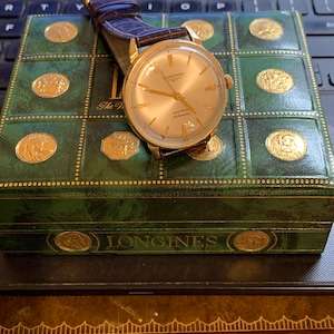 May include: A gold Longines Admiral Automatic wristwatch with a white face and black hands. The watch is sitting on a green box with gold accents and the word "Longines" printed on the front.