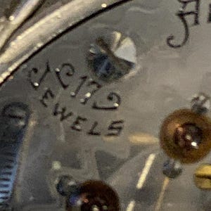 Op de afbeelding: Detailopname van een zilveren vintage horlogekast met de tekst "17 Jewels" in zwarte letters. Een kleine, fonkelende edelsteen en andere mechanische componenten zijn zichtbaar.