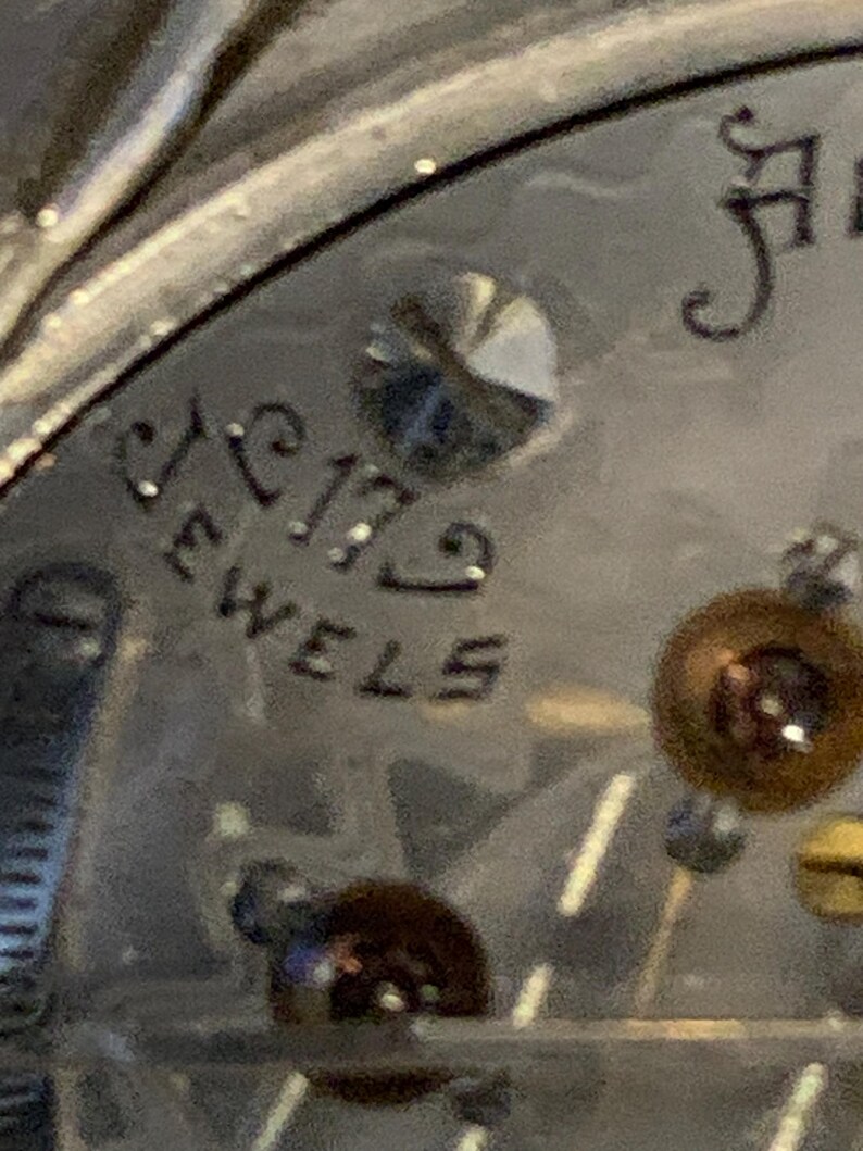 Op de afbeelding: Detailopname van een zilveren vintage horlogekast met de tekst "17 Jewels" in zwarte letters. Een kleine, fonkelende edelsteen en andere mechanische componenten zijn zichtbaar.