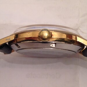 Puede incluir: Un reloj de pulsera dorado con una correa de cuero negro. El reloj tiene una esfera redonda con un bisel plateado y una corona dorada.