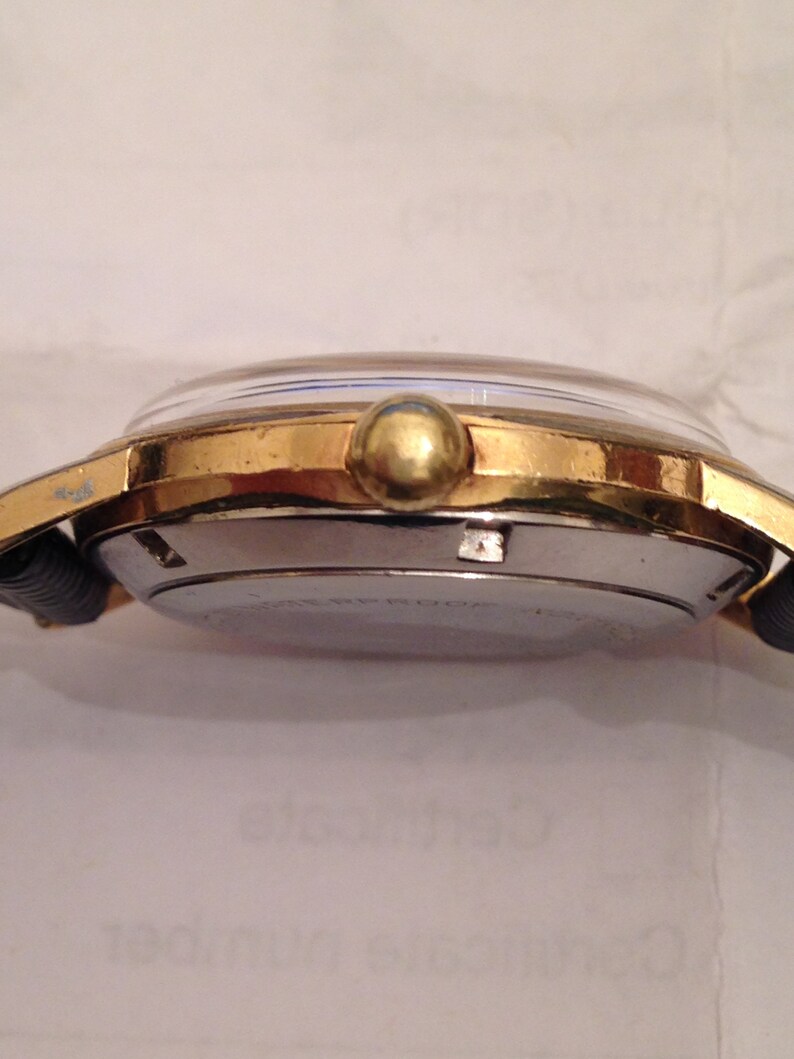 Puede incluir: Un reloj de pulsera dorado con una correa de cuero negro. El reloj tiene una esfera redonda con un bisel plateado y una corona dorada.