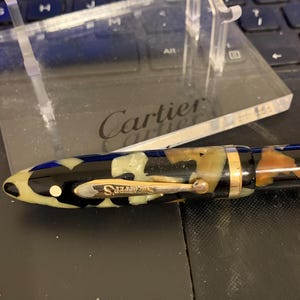 May include: A black and white patterned fountain pen with gold accents and the word "Sheaffer's" on the side. The pen is resting on a clear acrylic stand with the word "Cartier" engraved on it.