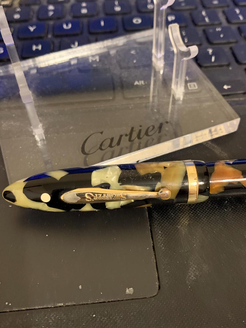 May include: A black and white patterned fountain pen with gold accents and the word "Sheaffer's" on the side. The pen is resting on a clear acrylic stand with the word "Cartier" engraved on it.