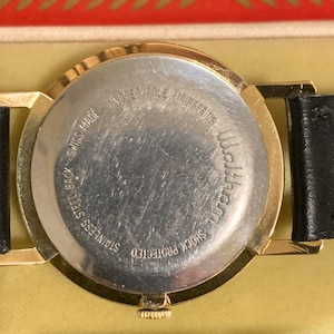 Op de afbeelding: De achterkant van een goudkleurig polshorloge met een zwarte leren band. De wijzerplaat van het horloge is zilverkleurig en heeft de tekst "Shock Protected", "Stainless Steel Back", "Swiss Made", "Unbreakable Mainspring" en "Waltham" erop gedrukt.