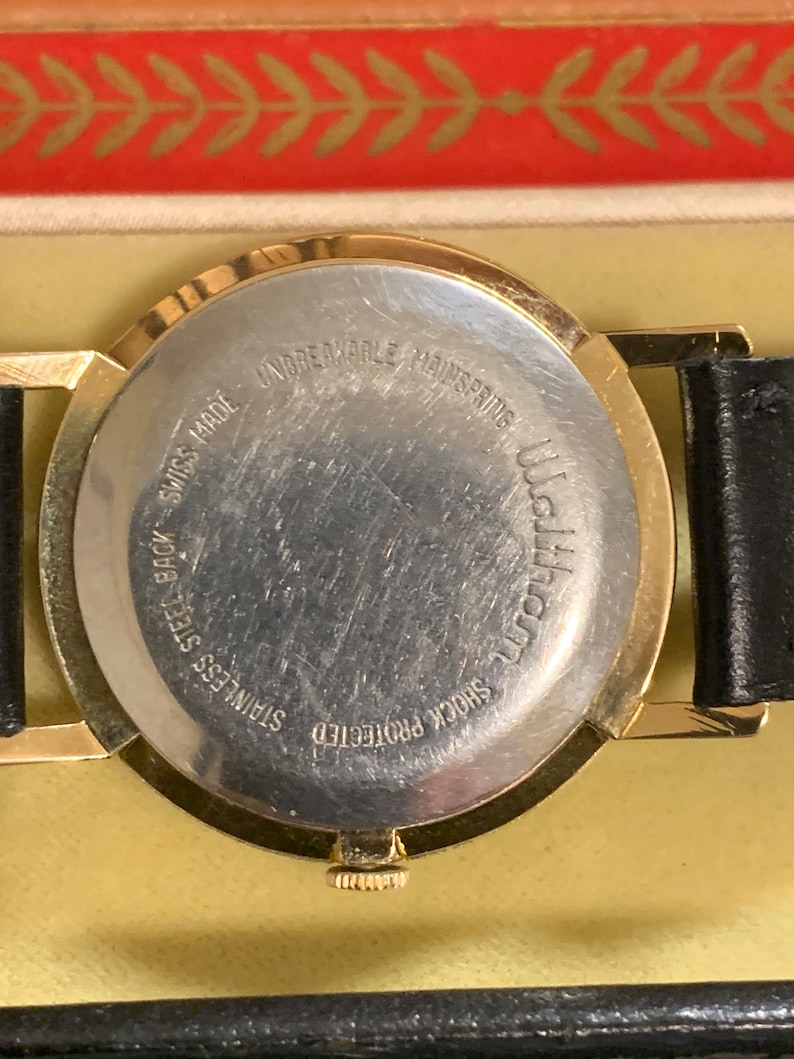 Op de afbeelding: De achterkant van een goudkleurig polshorloge met een zwarte leren band. De wijzerplaat van het horloge is zilverkleurig en heeft de tekst "Shock Protected", "Stainless Steel Back", "Swiss Made", "Unbreakable Mainspring" en "Waltham" erop gedrukt.