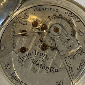 Op de afbeelding: Close-up van het uurwerk van een vintage Hamilton Watch Co. zakhorloge. Het zilverkleurige uurwerk toont de tekst "ADJUSTED", "179 JEWELS" en "Lancaster Pa.". De ingewikkelde tandwielen en componenten van het horloge zijn zichtbaar.
