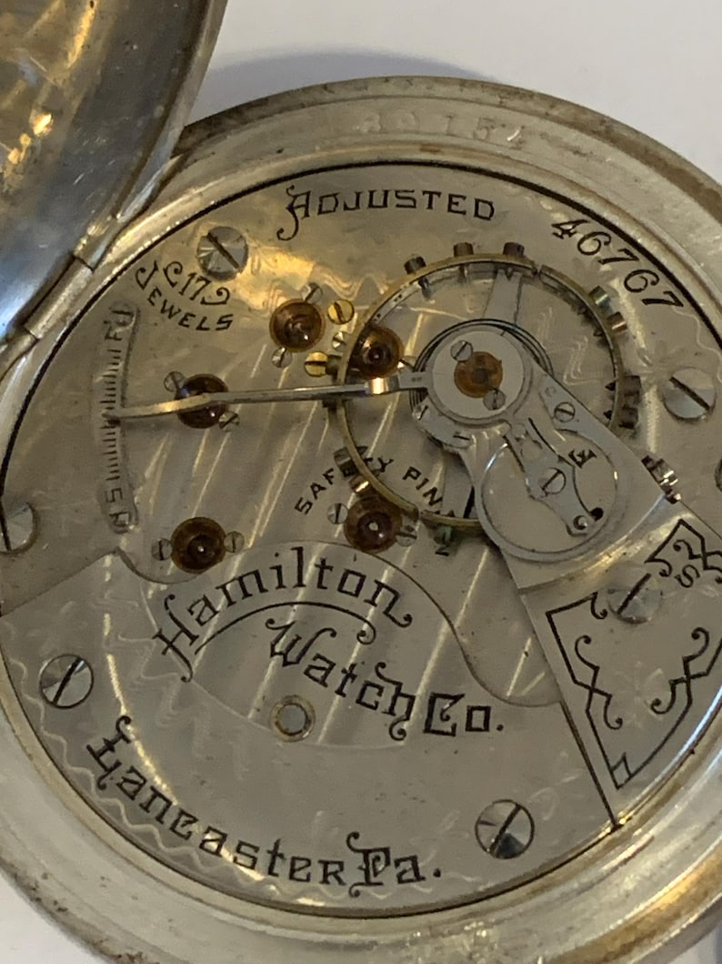 Op de afbeelding: Close-up van het uurwerk van een vintage Hamilton Watch Co. zakhorloge. Het zilverkleurige uurwerk toont de tekst "ADJUSTED", "179 JEWELS" en "Lancaster Pa.". De ingewikkelde tandwielen en componenten van het horloge zijn zichtbaar.