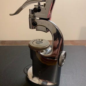 May include: A silver watch press with a lever and a round base. The press has a series of holes in the base for different sized watch cases.