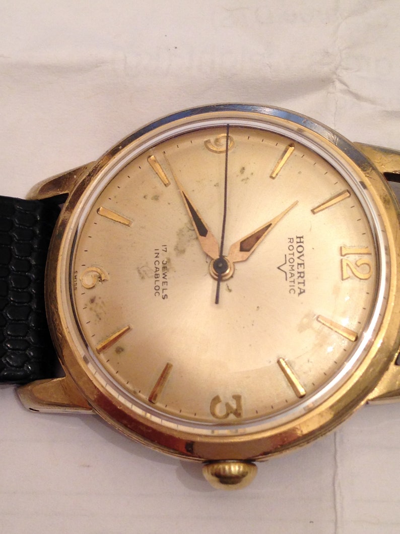 Puede incluir: Un reloj de pulsera dorado con una esfera blanca y agujas negras. La esfera del reloj tiene marcadores de hora romanos negros y una aguja de segundos negra. La esfera del reloj tambi&eacute;n tiene el texto "Hoverta Rotomatic 17 Jewels Incabloc" impreso en ella.