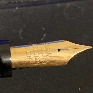 May include: Close-up of a gold-plated fountain pen nib with the inscription "WATERMAN'S IDEAL" and "14K" on the side.