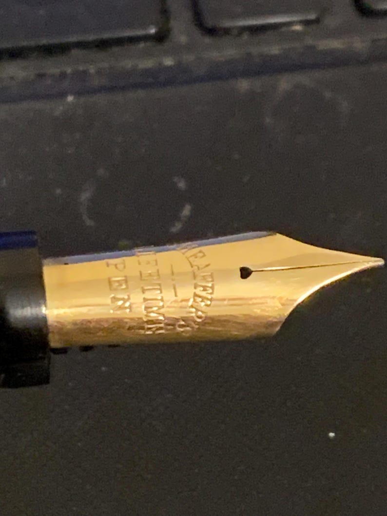 May include: Close-up of a gold-plated fountain pen nib with the inscription "WATERMAN'S IDEAL" and "14K" on the side.