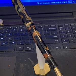 May include: A black and brown patterned fountain pen with a gold trim and a gold nib. The pen is on a gold stand.