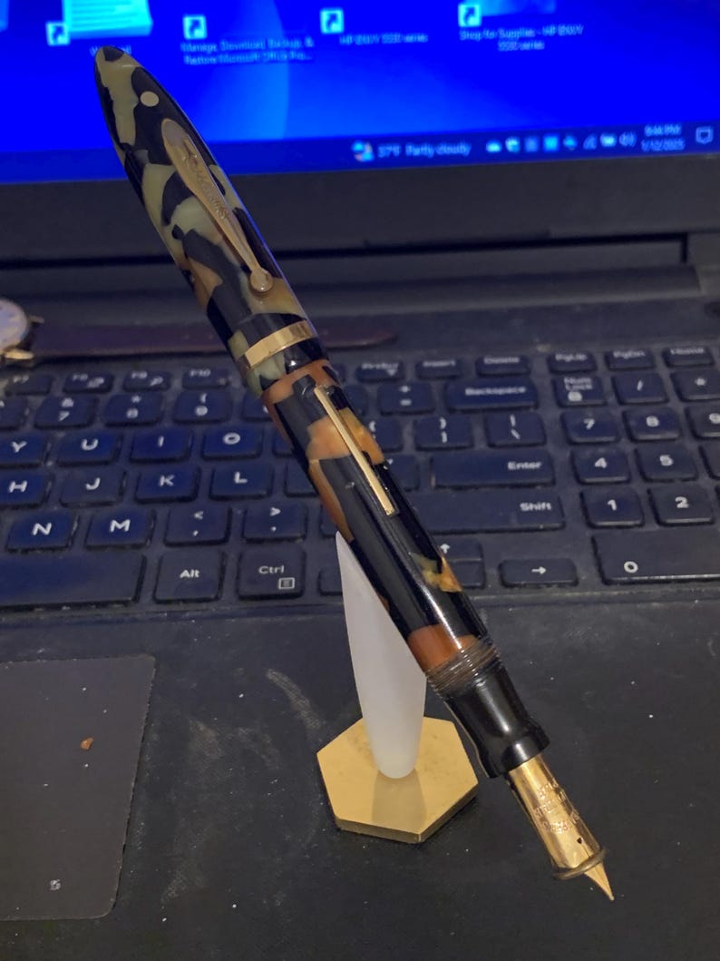 May include: A black and brown patterned fountain pen with a gold trim and a gold nib. The pen is on a gold stand.