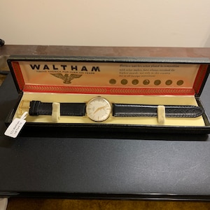 Op de afbeelding: Een gouden Waltham-horloge met een zwarte leren band in een zwarte doos. De doos heeft een rood en wit label met de tekst "Waltham Timing America for Over 100 Years".