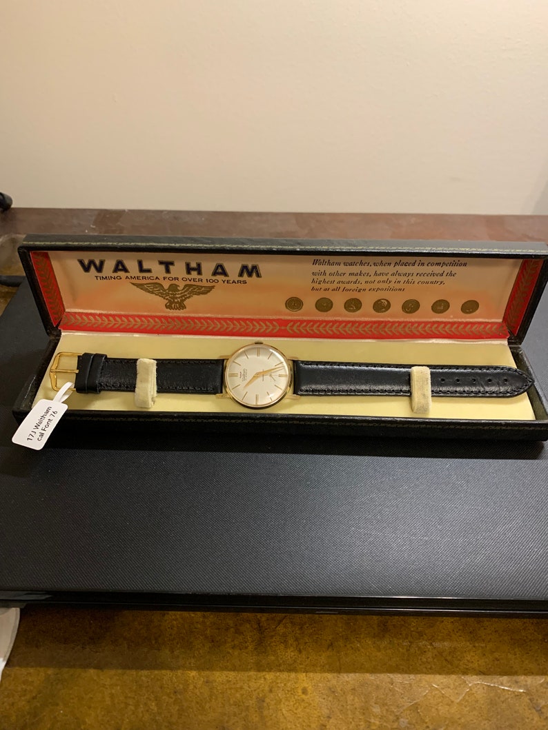 Op de afbeelding: Een gouden Waltham-horloge met een zwarte leren band in een zwarte doos. De doos heeft een rood en wit label met de tekst "Waltham Timing America for Over 100 Years".