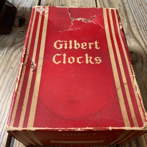 May include: A red cardboard box with white and gold stripes. The box has the text "Gilbert Clocks" printed on the front in white.
