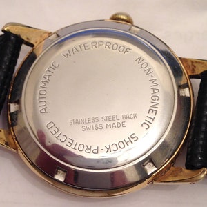 Puede incluir: La parte posterior de un reloj de pulsera dorado con una esfera plateada. El reloj est&aacute; etiquetado como "Automatic Waterproof Non-Magnetic Shock-Protected" y "Stainless Steel Back Swiss Made".
