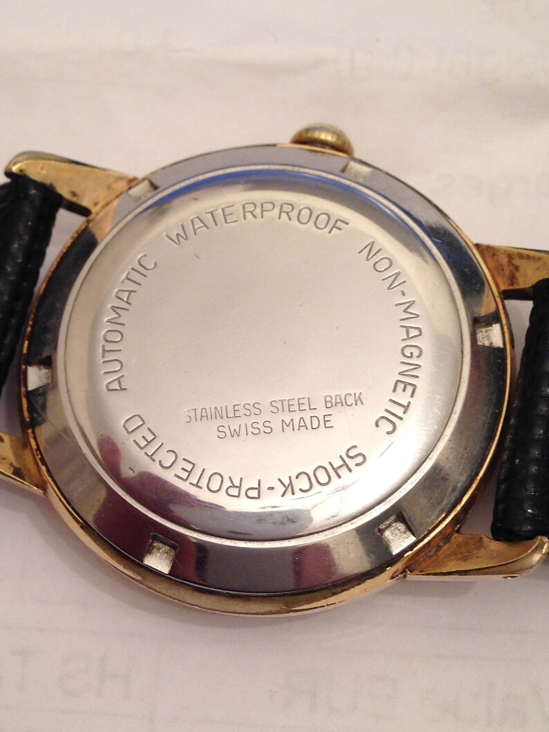 Puede incluir: La parte posterior de un reloj de pulsera dorado con una esfera plateada. El reloj est&aacute; etiquetado como "Automatic Waterproof Non-Magnetic Shock-Protected" y "Stainless Steel Back Swiss Made".