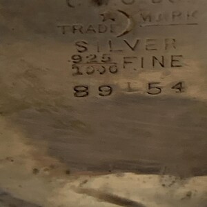Op de afbeelding: Close-up van een zilveren object met gestempelde tekst. De tekst bevat "CWC CO. MARK TRADE SILVER .925 FINE 1000" en het nummer "89154". Het object lijkt een stuk vintage zilverwerk te zijn.