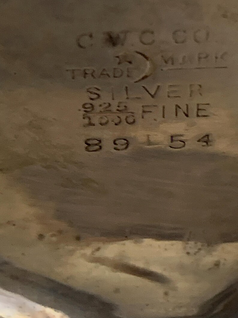Op de afbeelding: Close-up van een zilveren object met gestempelde tekst. De tekst bevat "CWC CO. MARK TRADE SILVER .925 FINE 1000" en het nummer "89154". Het object lijkt een stuk vintage zilverwerk te zijn.
