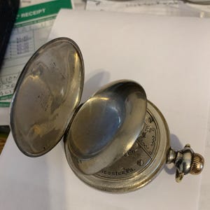 Op de afbeelding: Een open zilveren zakhorloge met een scharnierende klep. De wijzerplaat is zichtbaar, met ingewikkelde ontwerpen en de tekst "Lancaster Pa." De opwindknop heeft een goudkleurige accent.