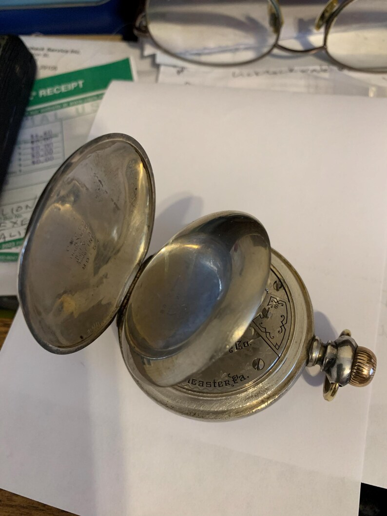 Op de afbeelding: Een open zilveren zakhorloge met een scharnierende klep. De wijzerplaat is zichtbaar, met ingewikkelde ontwerpen en de tekst "Lancaster Pa." De opwindknop heeft een goudkleurige accent.