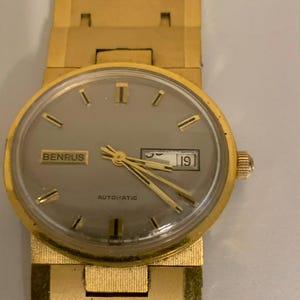 May include: A gold-toned Benrus automatic wristwatch with a gray face and a date display showing the number 19. The watch has gold hands and markers, and the band is also gold. The word "BENRUS" is printed on the watch face.